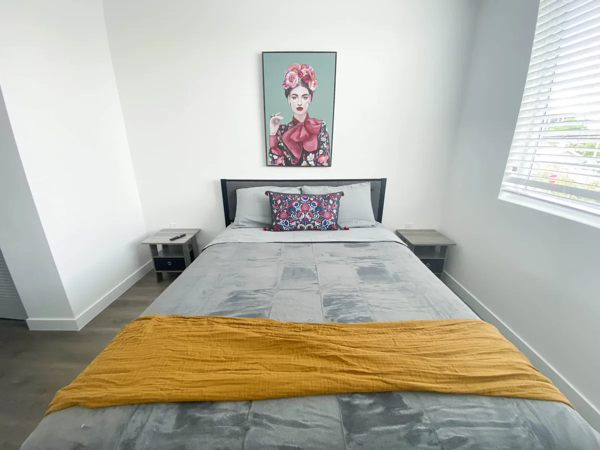 This image shows a serene bedroom featuring a comfortable bed with grey bedding, a colorful accent pillow, and a striking piece of artwork depicting a woman above the headboard. Natural light streams in, creating an inviting atmosphere ideal for unwinding or enjoying a quiet morning.