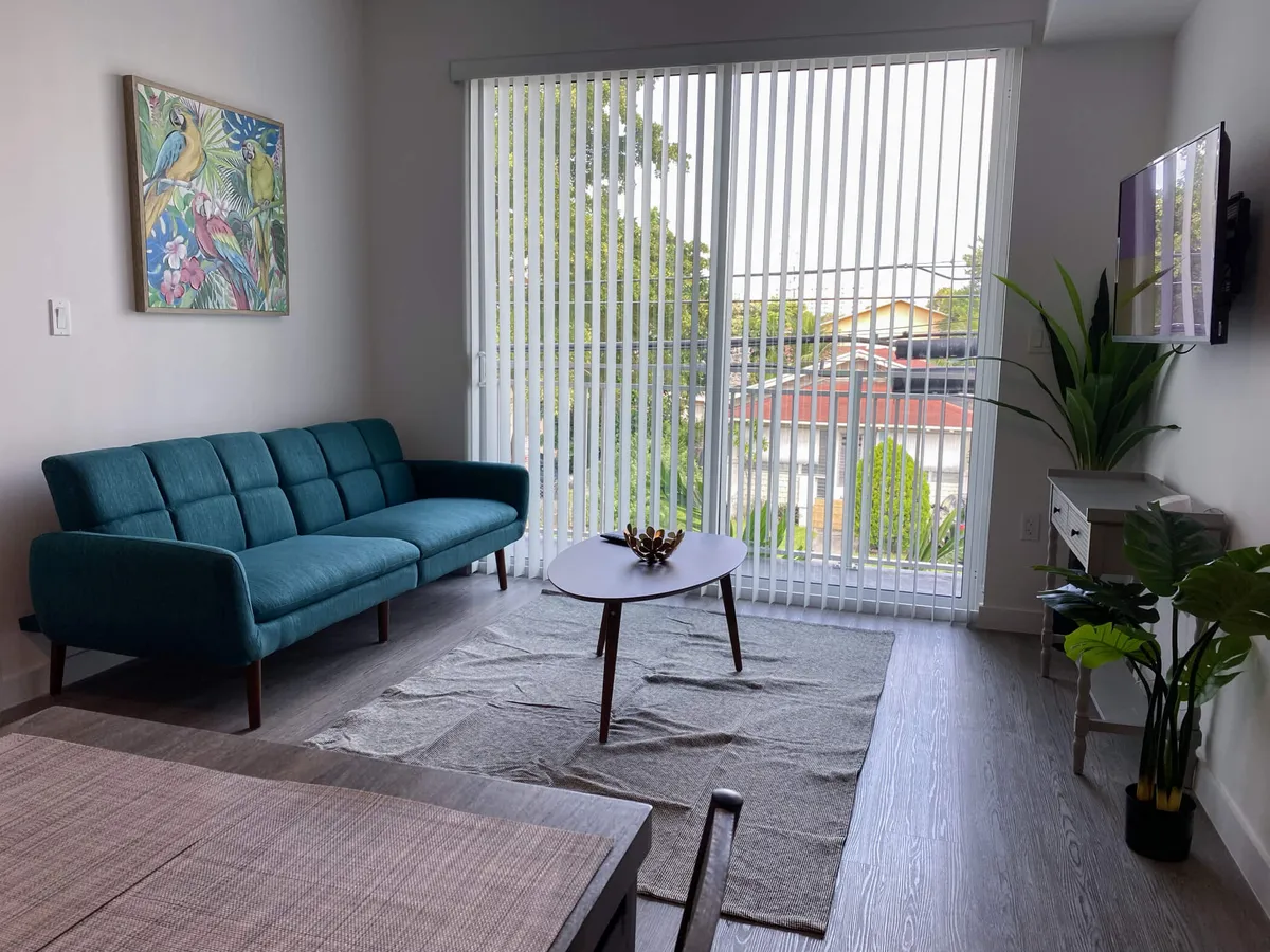 The hero image showcases a bright, airy living room with a chic teal blue mid-century modern sofa, a sleek oval wooden coffee table, and a wall-mounted flat screen TV. Natural light floods the space through a large sliding glass door with vertical blinds, creating a warm, inviting atmosphere ideal for relaxing or socializing.