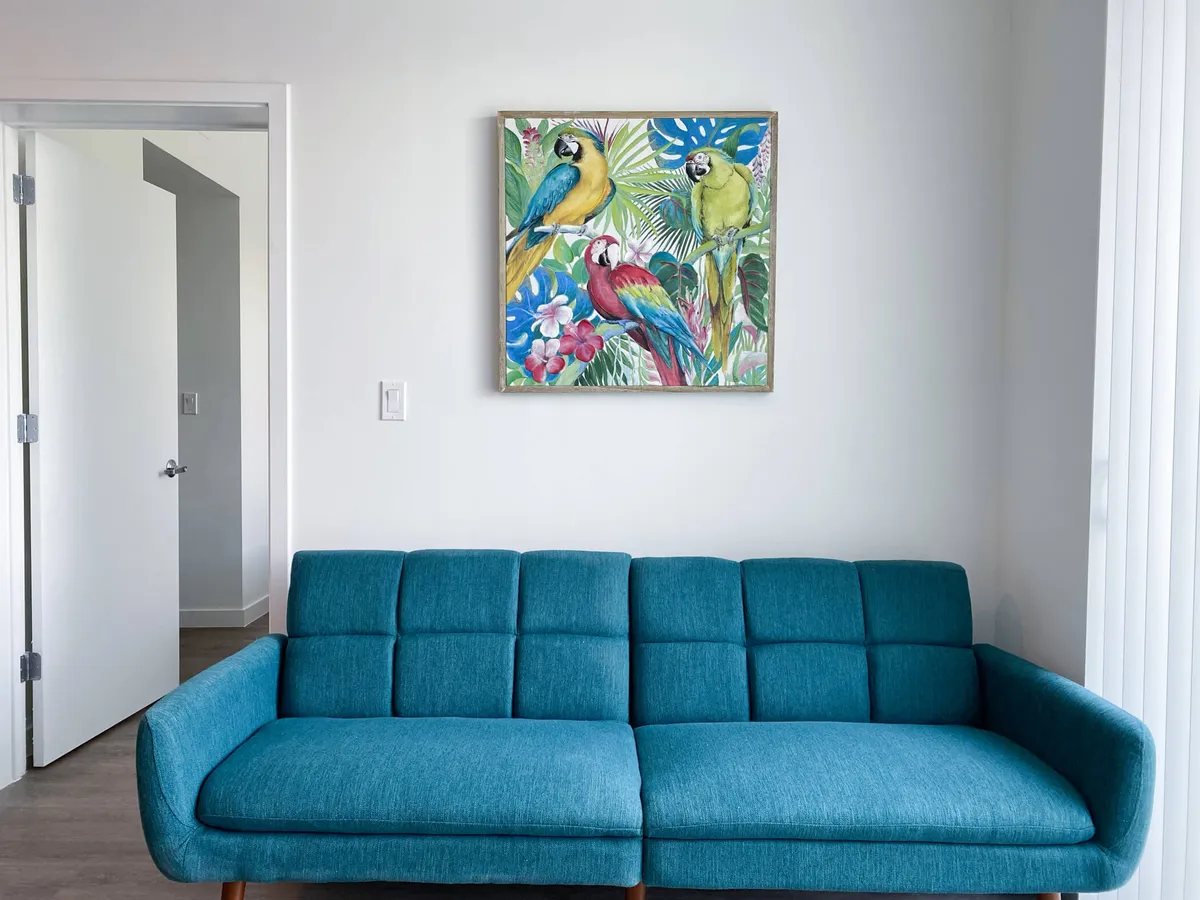 A welcoming living room featuring a blue upholstered sofa beneath vibrant parrot artwork. The open white door leads to another room, subtly hinting at the apartment's open layout and easy flow between spaces. The setting is clean, cozy, and inviting, ideal for both relaxing and spending time with a pet.
