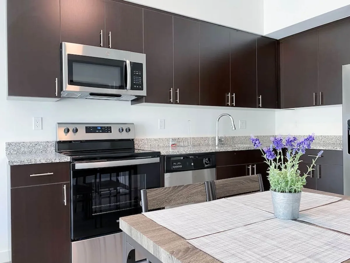 This image displays a modern, open kitchen with stainless steel oven and microwave, dark wood cabinetry, and granite countertops. The scene includes a dining table set with a potted lavender centerpiece, emphasizing both the cooking and gathering aspects. Modern appliances and full kitchen setup are clearly visible, supporting the section's emphasis on a fully equipped kitchen and dining area.