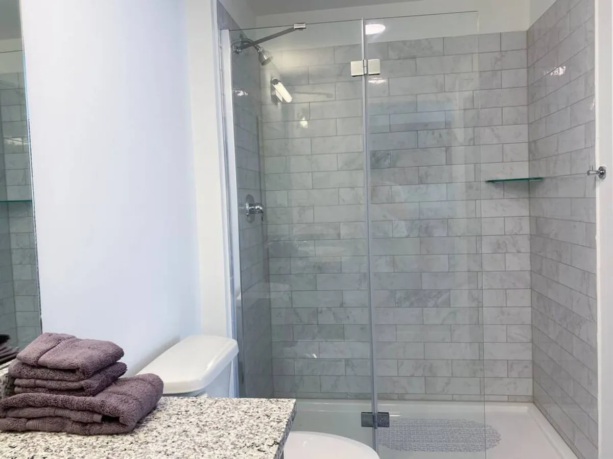 A pristine bathroom with a glass shower enclosure, marble tile walls, a granite countertop with neatly folded towels, and a white toilet. The setting is fresh, calming, and well-stocked, hinting at thoughtful amenities provided for guests.
