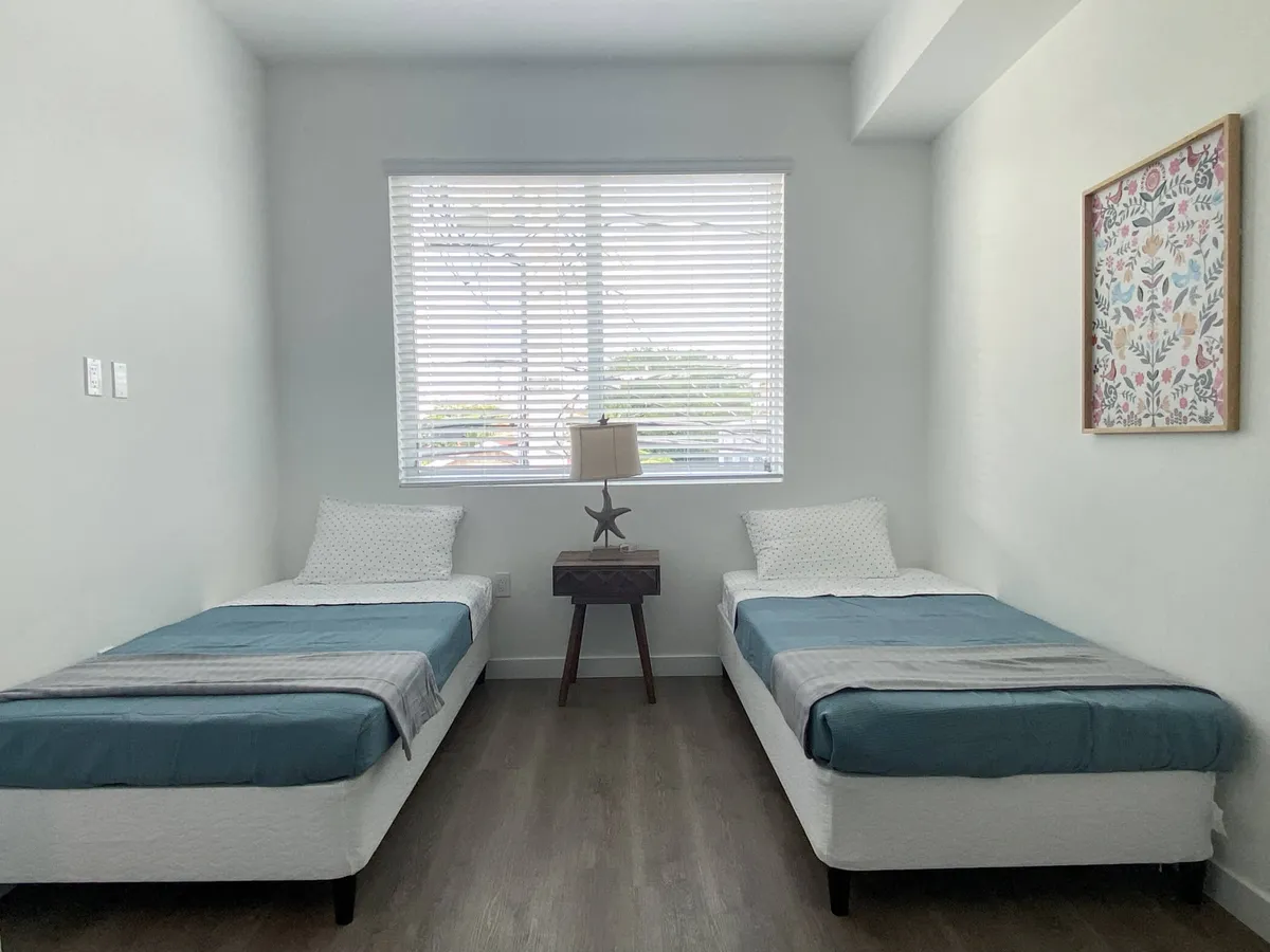 A tranquil twin bedroom with two single beds outfitted in blue and gray bedding, separated by a wooden nightstand topped with a starfish-shaped lamp. A large window brings in daylight, and colorful framed artwork adds a cheerful touch to the uncluttered, peaceful space.