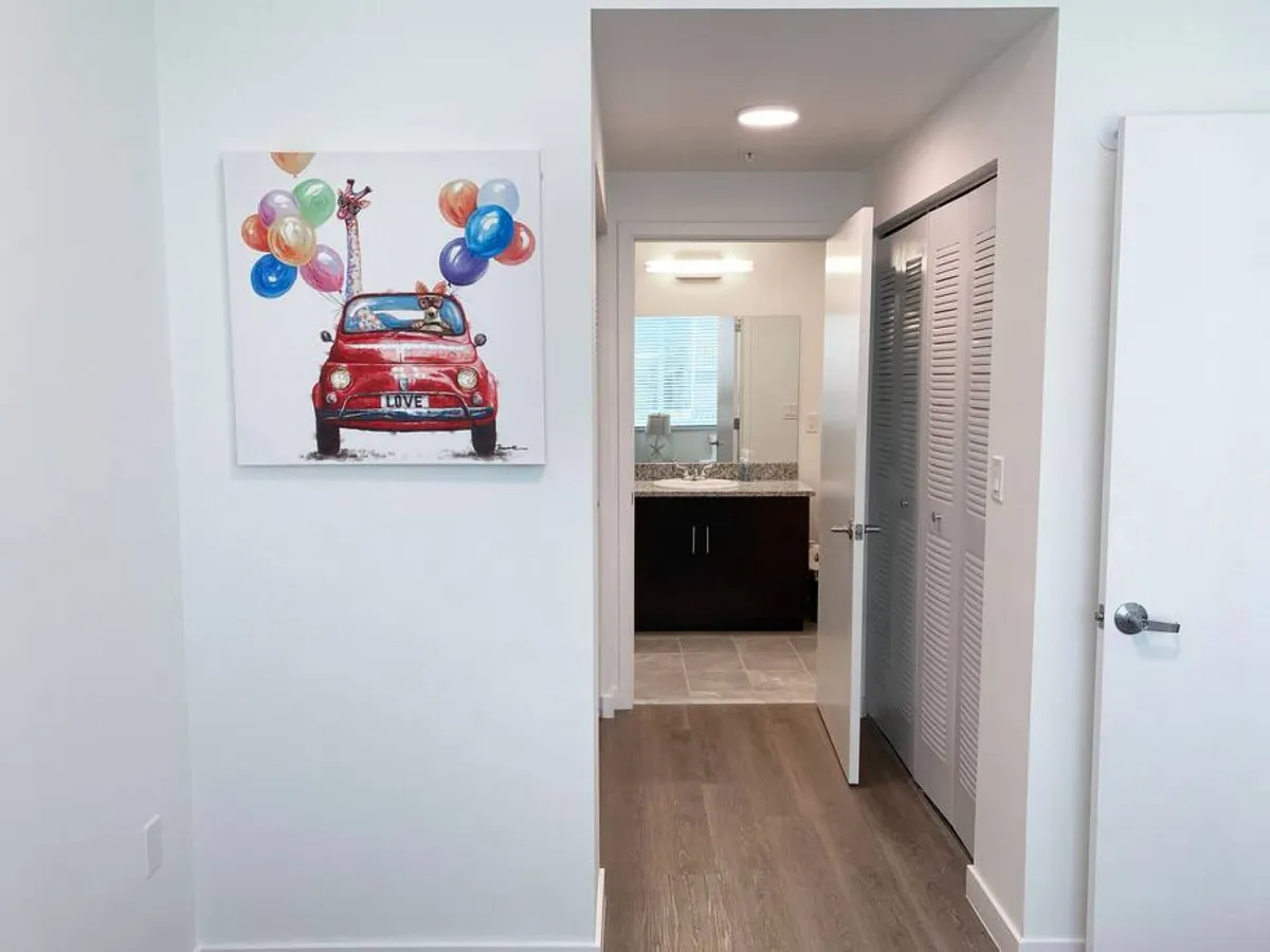 A cheerful, inviting hallway featuring playful artwork of a red car with balloons and animals. The scene includes an open doorway to a bathroom with granite countertops, a closet with louvered doors, and another open white door leading to an adjacent area. The decor infuses character and warmth while illustrating the ease of movement and access to daily necessities.