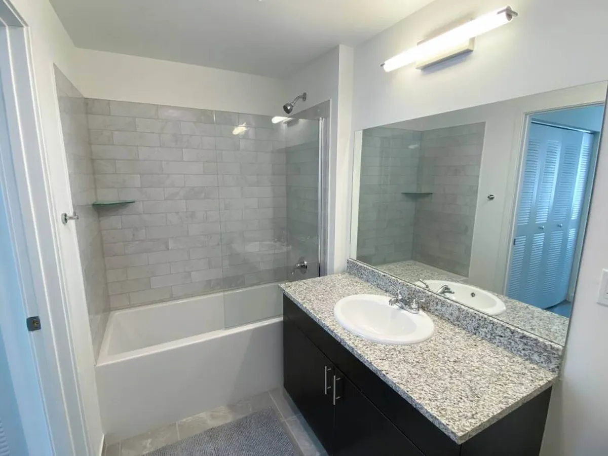 A modern full bathroom featuring a granite countertop with built-in sink, a large mirror above, and a bathtub with a glass shower screen. Subtle tile accents and bright lighting create a fresh, practical space for daily self-care.